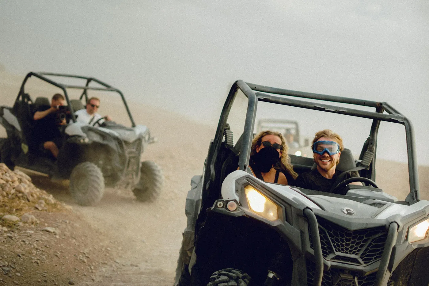 Deelnemers rijden lachend in buggy's door het stof, met een bril op tegen het zand.